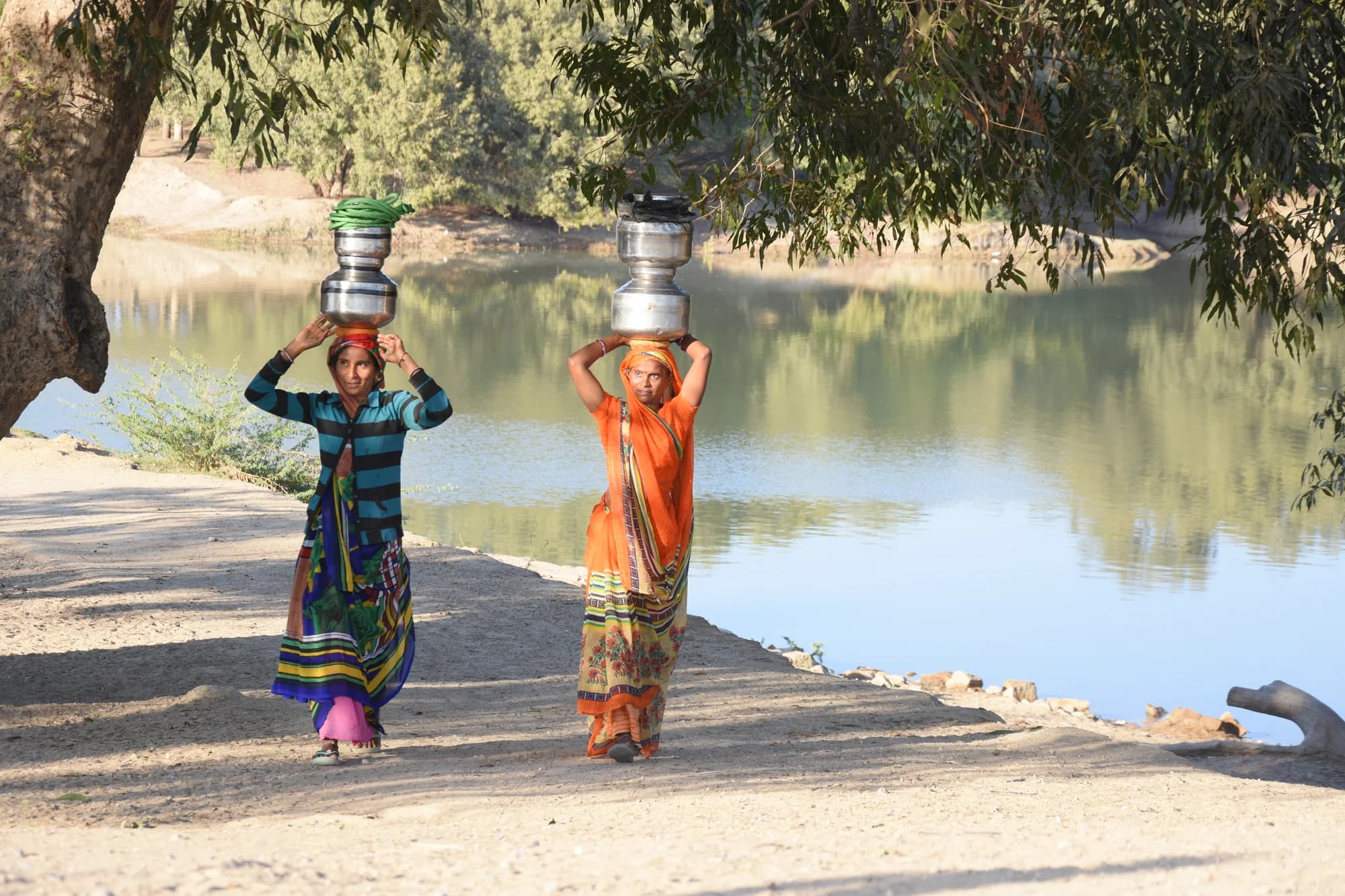 Two village women carrying water pots on their heads beside a lake